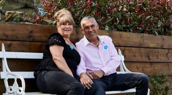 A woman and man sit on a garden bench smiling, wearing Alzheimer's Society Forget Me Not badges